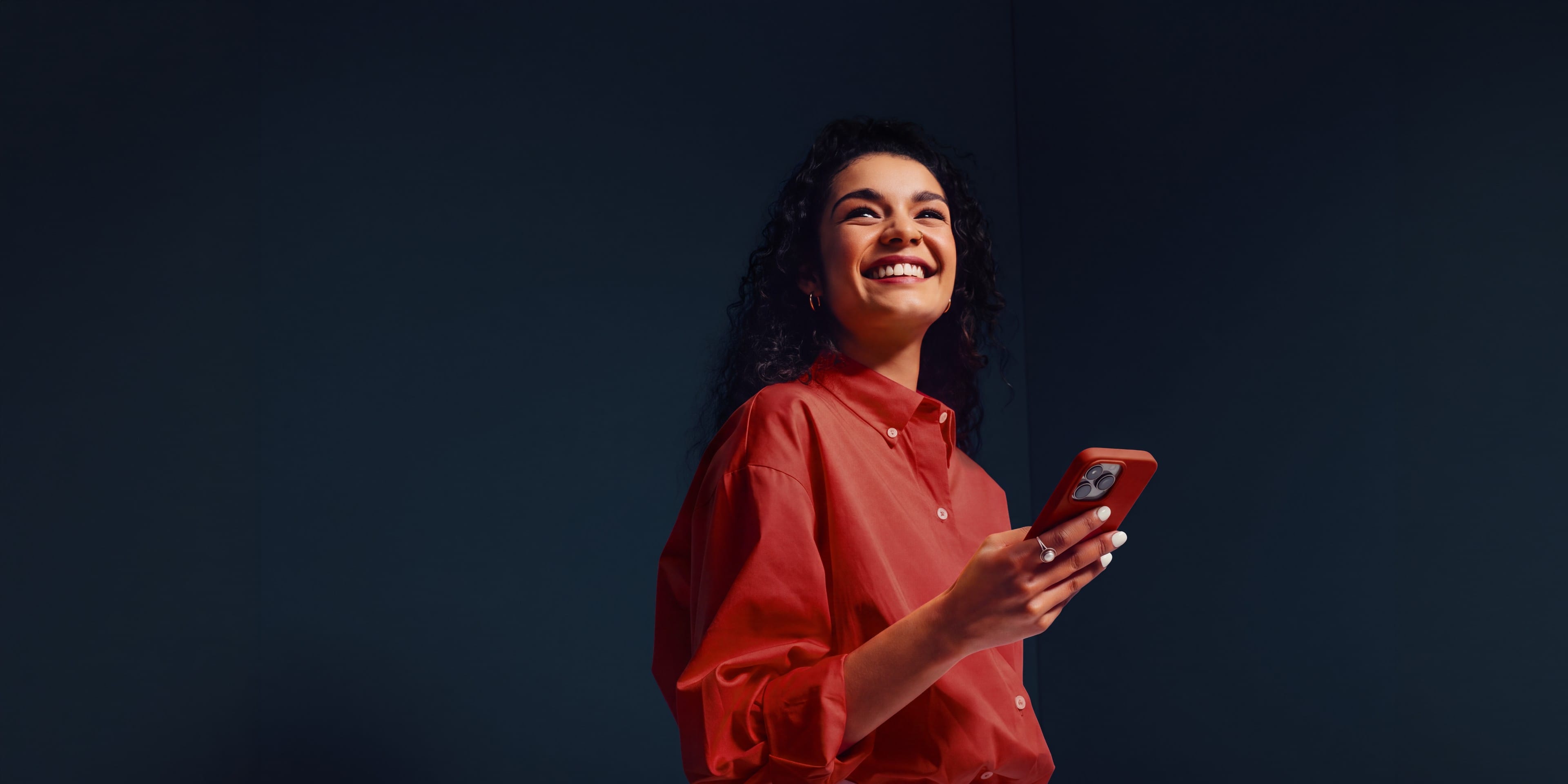 A woman in a red shirt laughing with her phone in hand against a deep teal wall