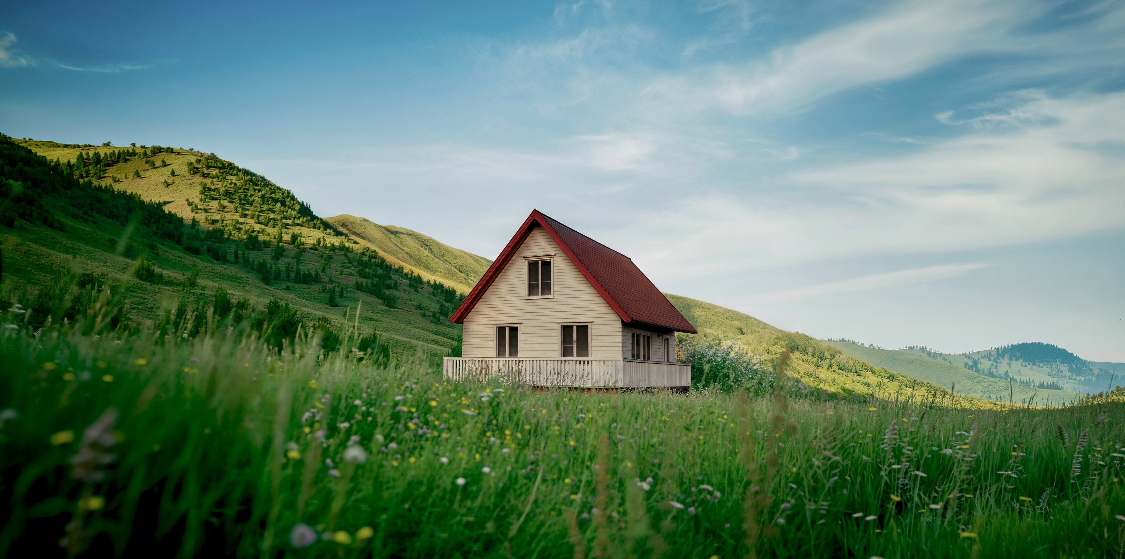 A white house with a red roof in a meadow of tall grass, green hills rising behind it