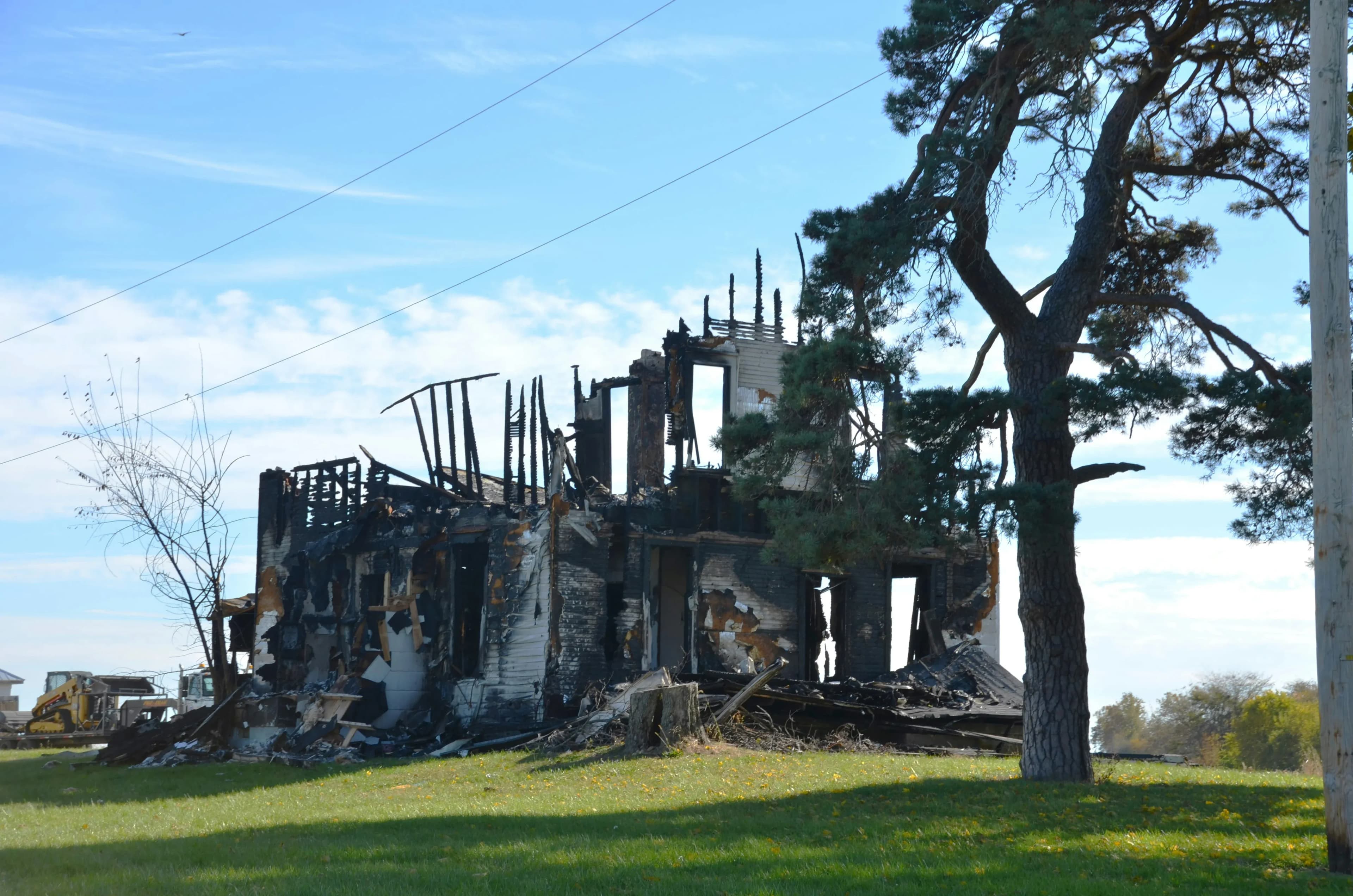 The burned-out frame of a home under a blue sky, with a large tree in the yard.