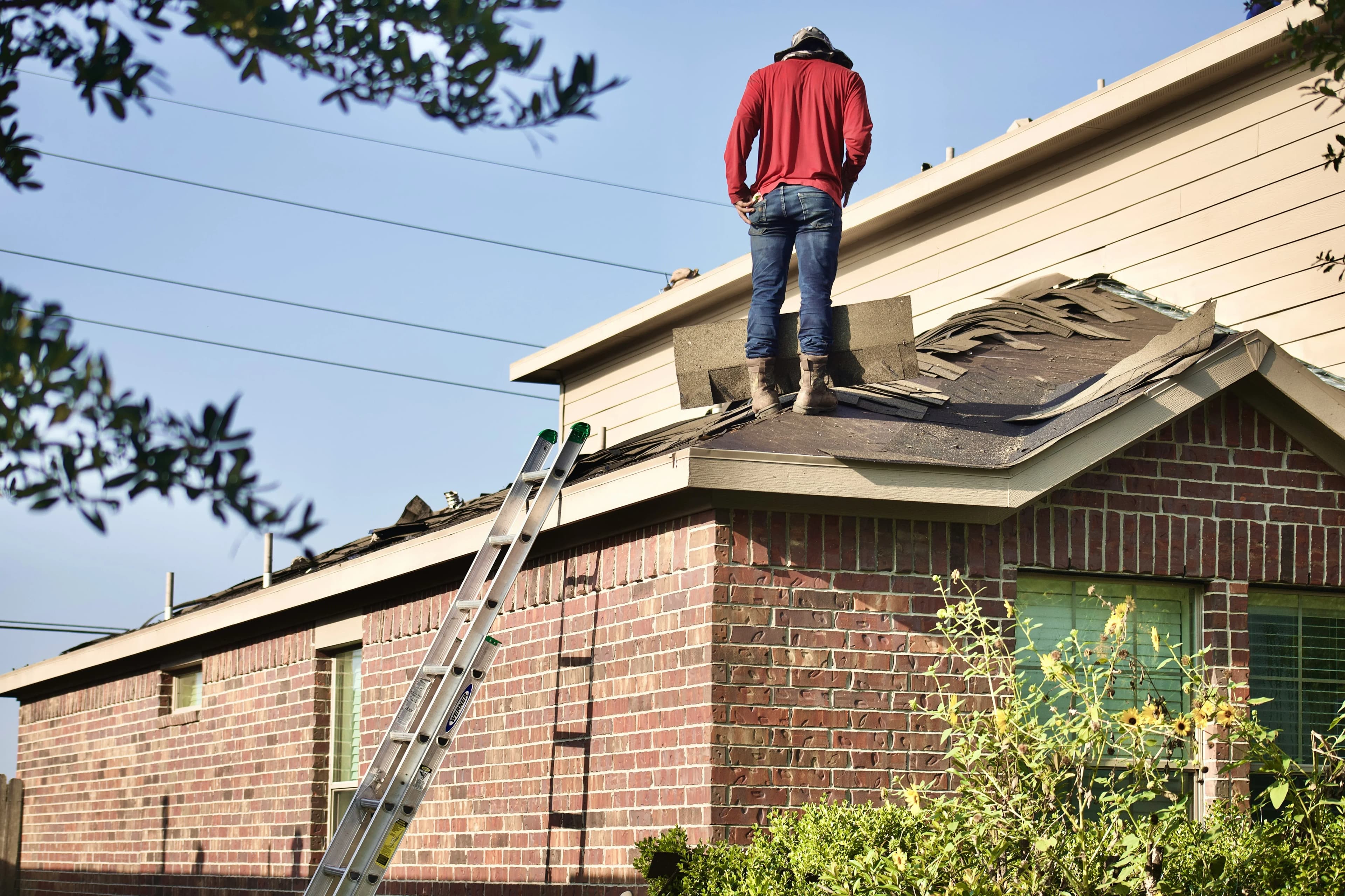 A roofer stands on the roof of a home, looking at wind storm damage and missing shingles.