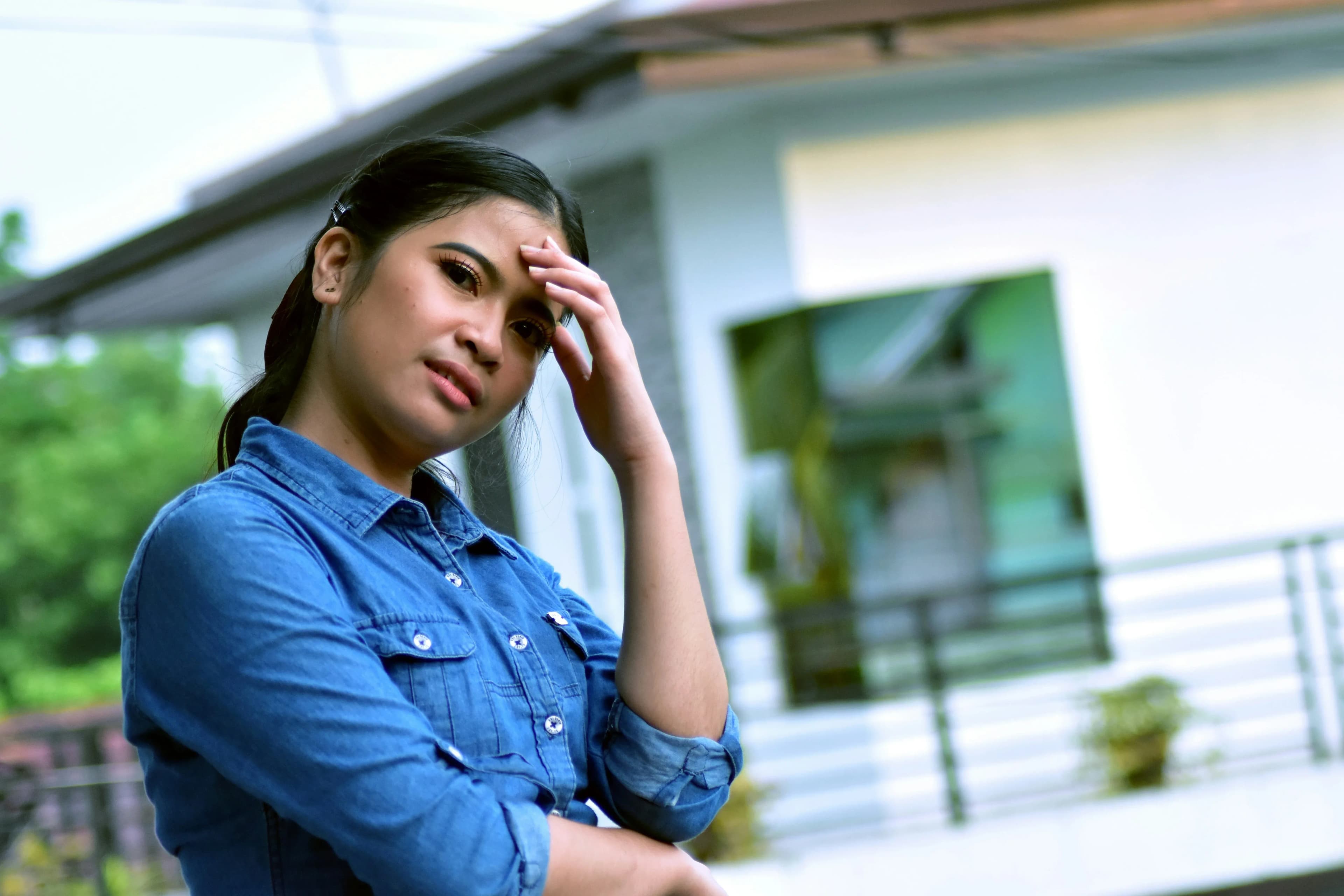 A woman standing in front of her home looks regretful and concerned, with her hand to her forehead.