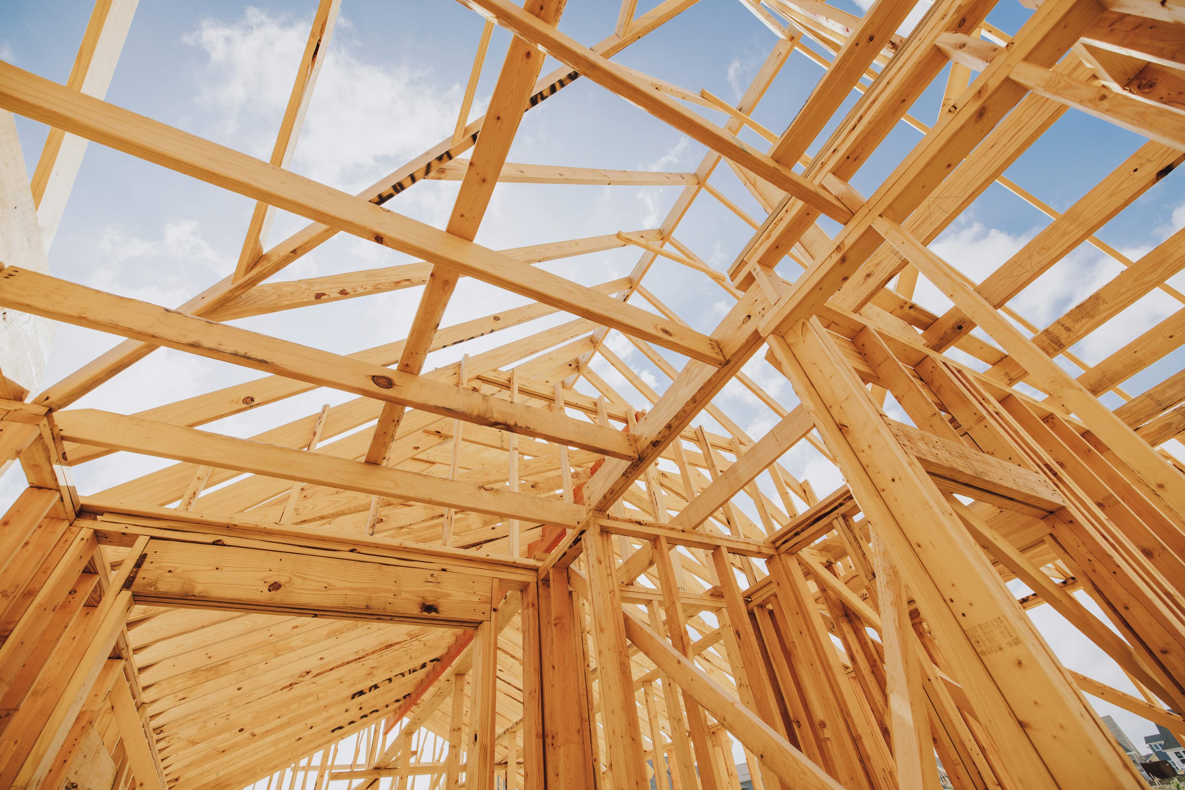 A new wooden frame home in the process of being built is shown from inside, with a cloudy sky visible above.